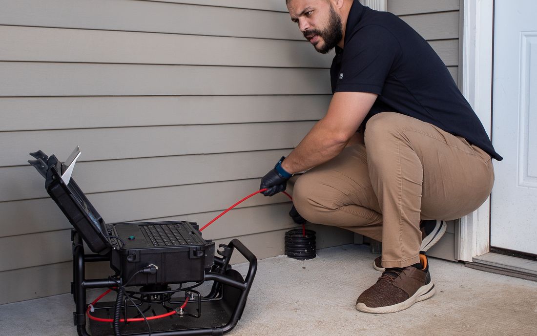 A technician feeding a sewer camera into an outdoor cleanout, with the camera reel and monitor beside him