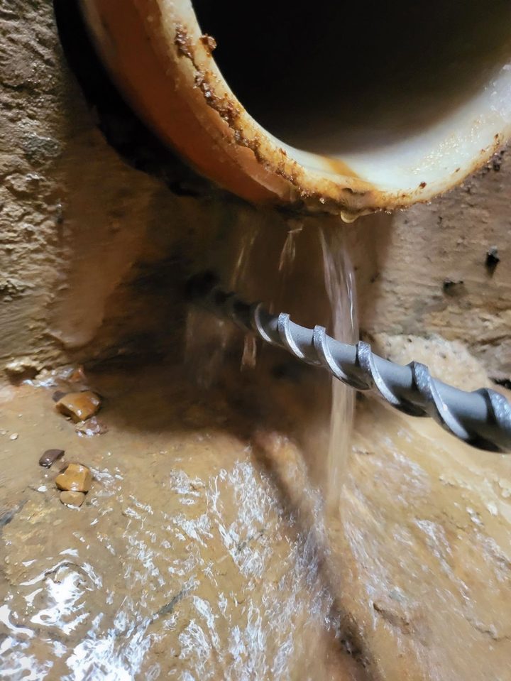 A drill bit going into the wall beside a pipe as water pours out